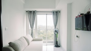 A minimalist living room with a white sofa, a standing fan, and a large window with curtains. - Wonderful 1Br Casa De Parco Apartment Near Ice Bsd (South Tangerang)