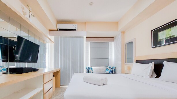 A modern hotel room with a large bed, a flat-screen TV, a desk with a computer, and a large window with blinds. - Cozy And Simply Studio Apartment At Urban Heights Residences (South Tangerang)