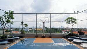 Una cancha de baloncesto al aire libre con aro, rodeada por una valla metálica, con un paisaje urbano de fondo.