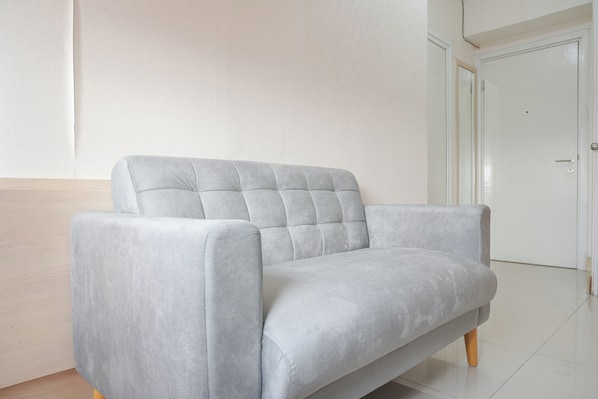 Un canapé gris clair, doté de pieds en bois, se trouve dans une pièce aux murs blancs, avec une porte fermée en arrière-plan.