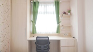 A small, well-lit room with a desk, a chair, a window with green curtains, a shelf with decorative items, and a wall-mounted air conditioner. - Good Deal 1Br Green Pramuka City Near Mall Apartment (Jakarta)