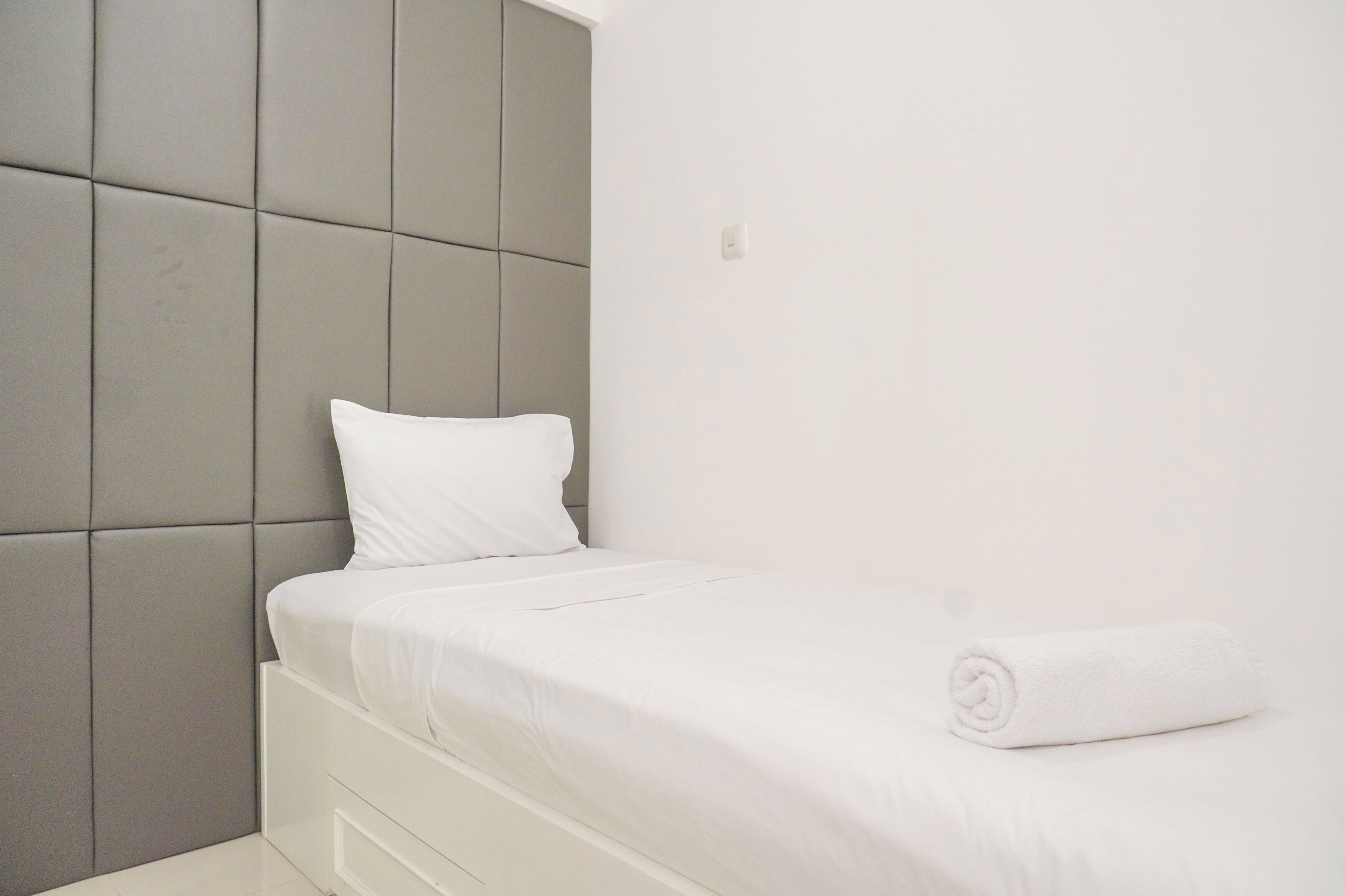 Un dormitorio moderno con un cabecero acolchado gris, una cama bien hecha con ropa blanca y una toalla blanca enrollada sobre la cama.
