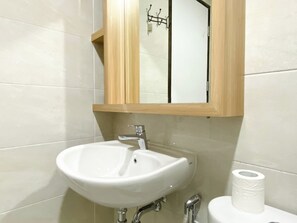 A bathroom with a white sink, a wooden medicine cabinet above it, and a toilet with a roll of toilet paper. - Stunning And Homey 2Br Apartment Mustika Golf Residence (Cikarang)
