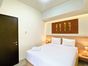 A hotel room with a bed, a wooden headboard, a wall-mounted artwork, a bedside table with a lamp, and a door. - Stunning And Homey 2Br Apartment Mustika Golf Residence (Cikarang)