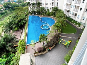Uma piscina com formato singular, rodeada por vegetação exuberante e um edifício de vários andares ao fundo.