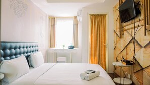 Una habitación de hotel con un cabecero capitonado, una cama con ropa de cama blanca, un escritorio pequeño con una silla, un televisor montado en una pared de madera y una ventana con cortinas.