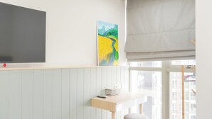 Une pièce dotée d’un grand téléviseur à écran plat, d’un tableau accroché au mur, d’un petit bureau avec une chaise et d’une fenêtre donnant sur des immeubles.