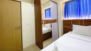 A hotel room with a wooden wardrobe, a bed with white linens, and a window with blue curtains. - Spacious And Comfy 2Br With Extra Room At Meikarta Apartment (Cikarang)