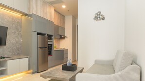 A modern kitchen with grey cabinets, a built-in oven, and a flat-screen TV. There is a grey sofa with a houndstooth pattern and a small table with a water bottle on it. - Gorgeous And Comfortable 1Br Sudirman Suites Apartment (Jakarta)