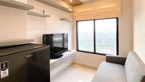 A modern living room with a flat-screen TV, a grey sofa, and a large window offering a city view. - Cozy And Comfort 2Br At Pollux Chadstone Apartment (Cikarang)