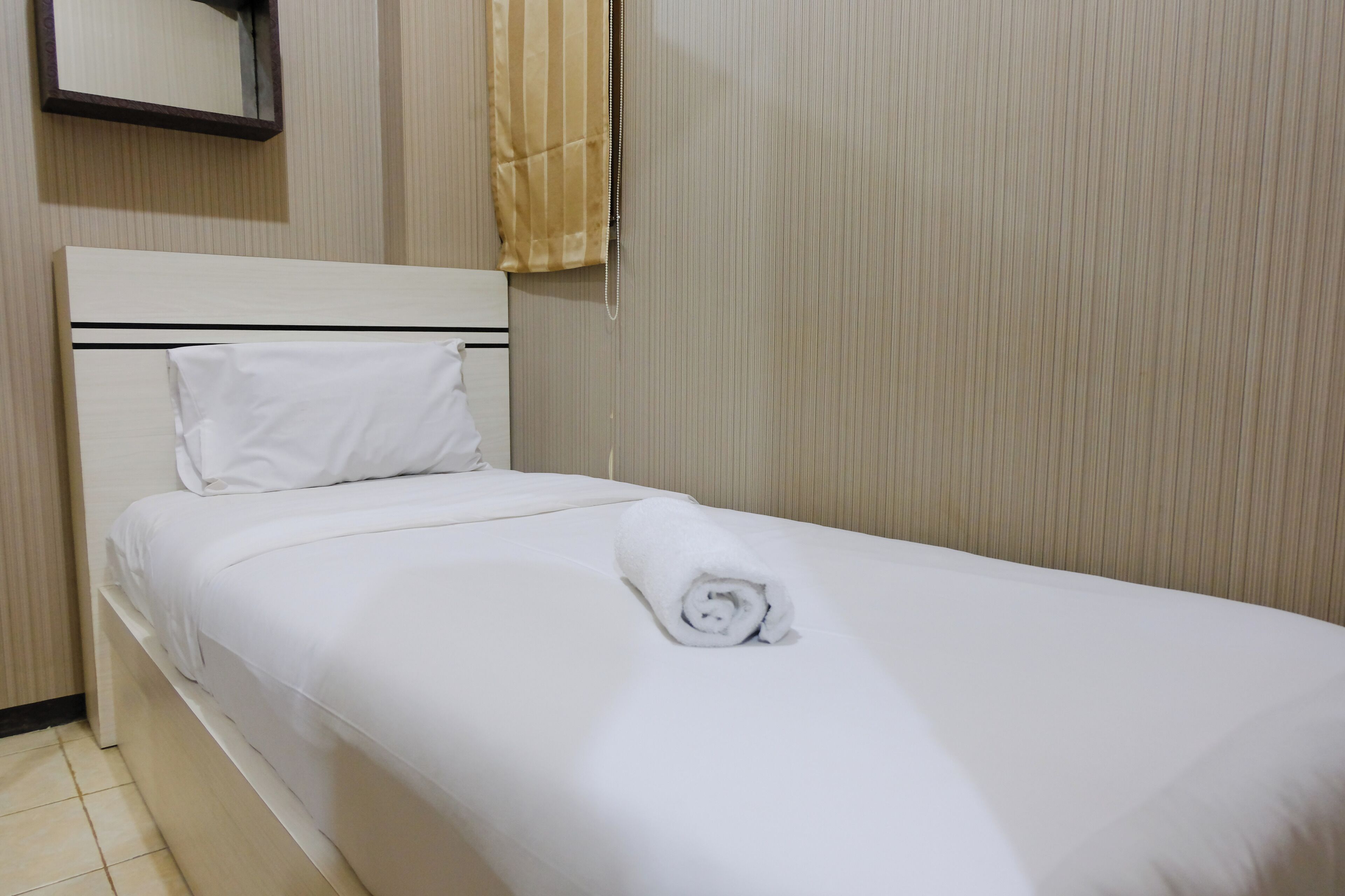 A neatly made bed with white linens and a rolled towel, a wooden headboard, and a wall-mounted mirror.
