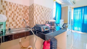 A compact kitchen with a sink, stove, and kettle, a water dispenser, and a window with blue curtains. - Good Choice Studio At Margonda Residence 5 Apartment (Depok)