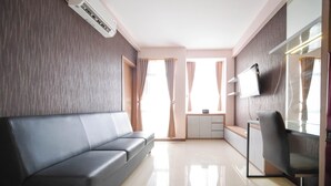 A modern hotel room with a grey sofa, a desk with a chair, a flat-screen TV, and an air conditioning unit. - Spacious 2Br Combine Unit At Bale Hinggil Apartment (Surabaya)