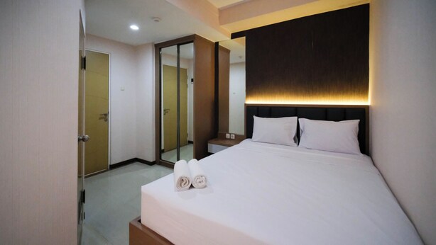 A hotel room with a neatly made bed, a bedside table, a mirror, and a door leading to another room. - Spacious 2Br Combine Unit At Bale Hinggil Apartment (Surabaya)