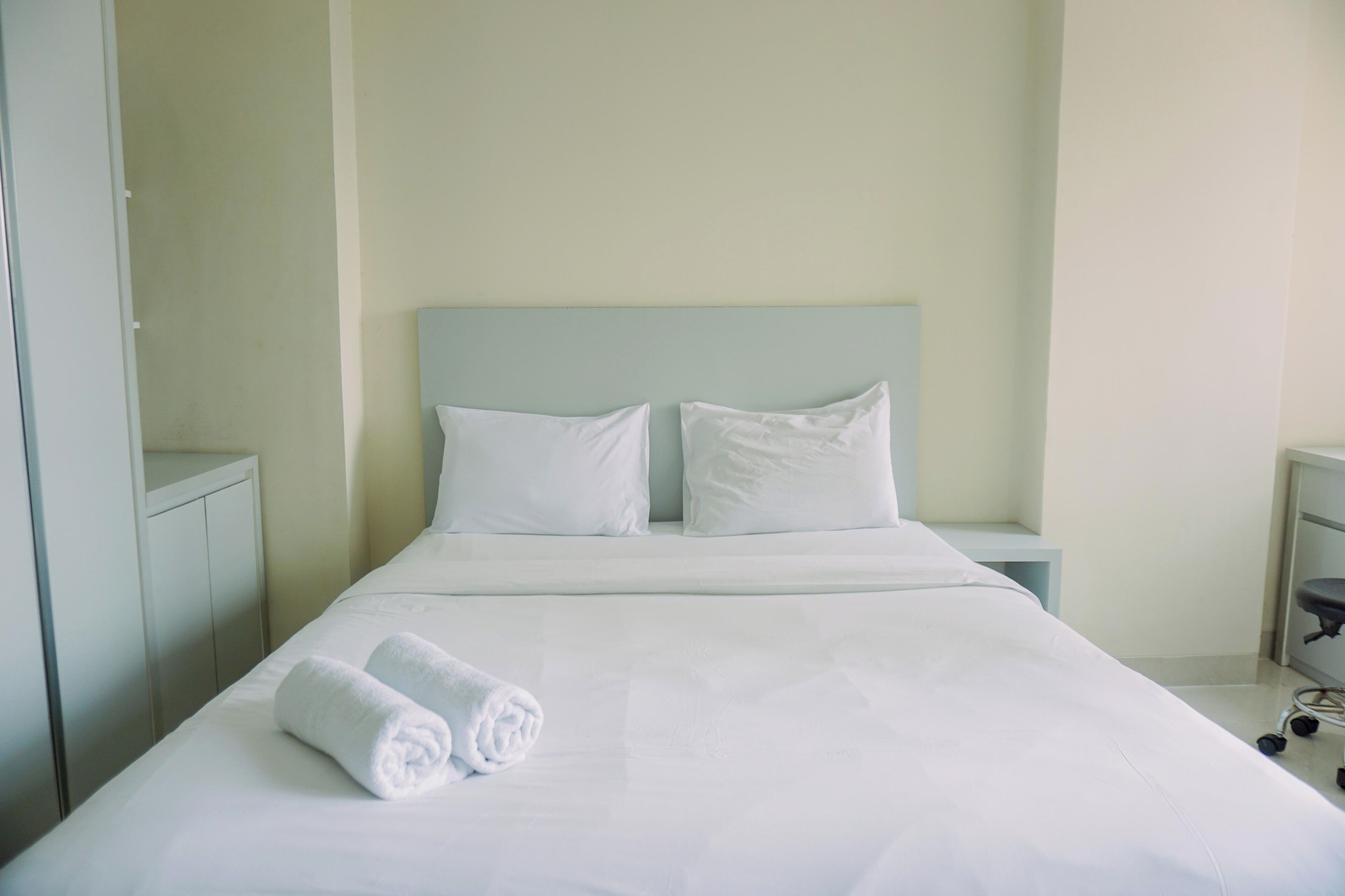 A neatly made bed with white linens and two pillows, a bedside table with a lamp, and a small desk with a chair.