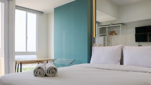 A neatly made bed with rolled towels, a window with a view, a white table, and a blue accent wall. - Best Deal And Comfort Studio Tokyo Riverside Pik 2 Apartment (Teluknaga)