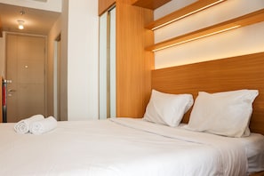 A hotel room with a wooden headboard, a neatly made bed with white linens, and a visible door to the left. - Comfy And Tidy Studio At Tokyo Riverside Apartment Pik 2 (Teluknaga)