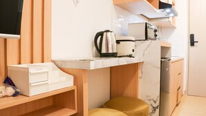 Une cuisine compacte équipée d’un plan de travail, d’une bouilloire, d’une machine à café et d’un four à micro-ondes. On y trouve une étagère en bois, une boîte blanche et un tabouret jaune.
