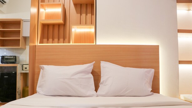 Un lit bien fait, avec du linge de lit blanc et des oreillers, une tête de lit en bois et une étagère intégrée au-dessus du lit.