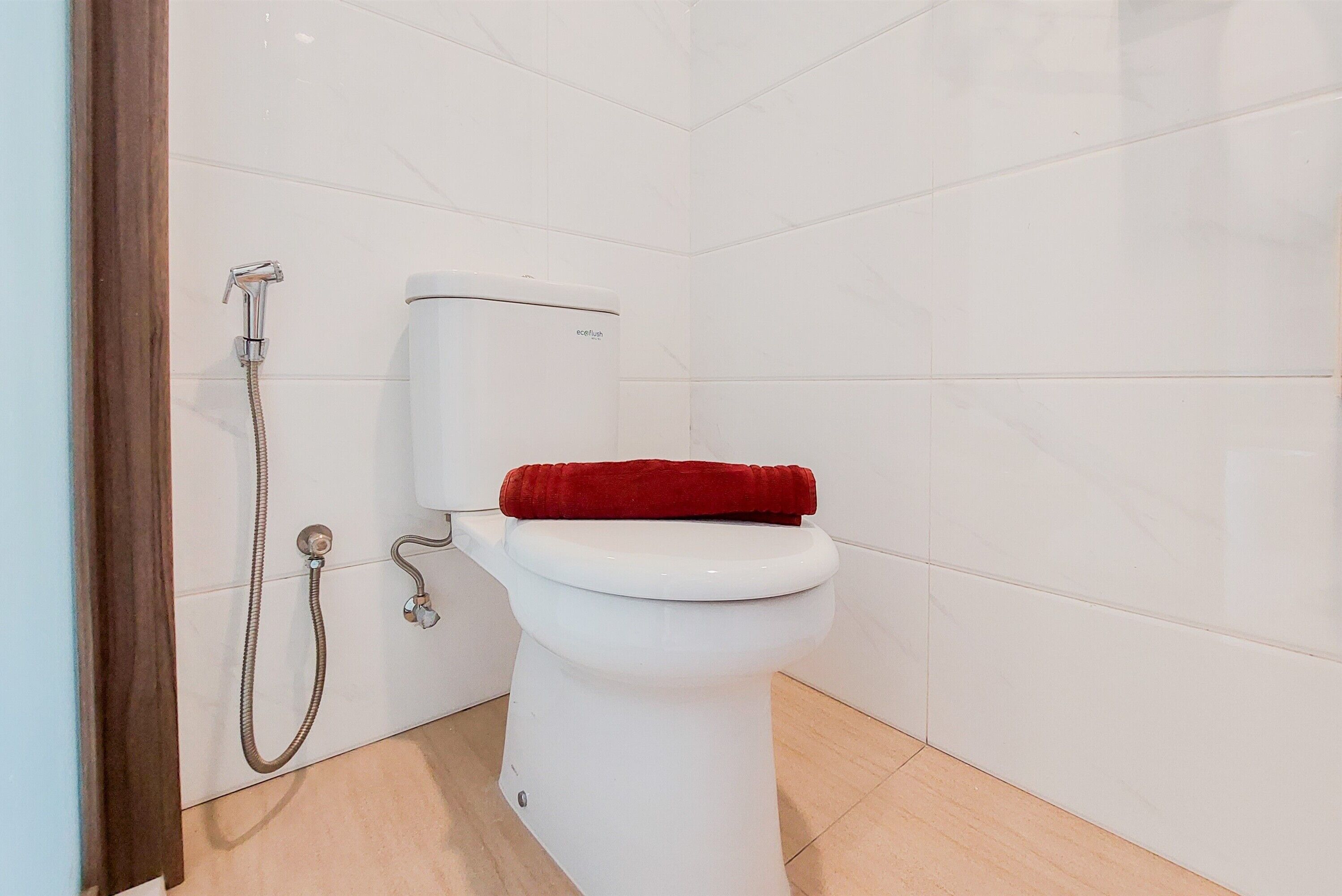 A white toilet with a red towel on top, a showerhead, and a wall-mounted flush handle.