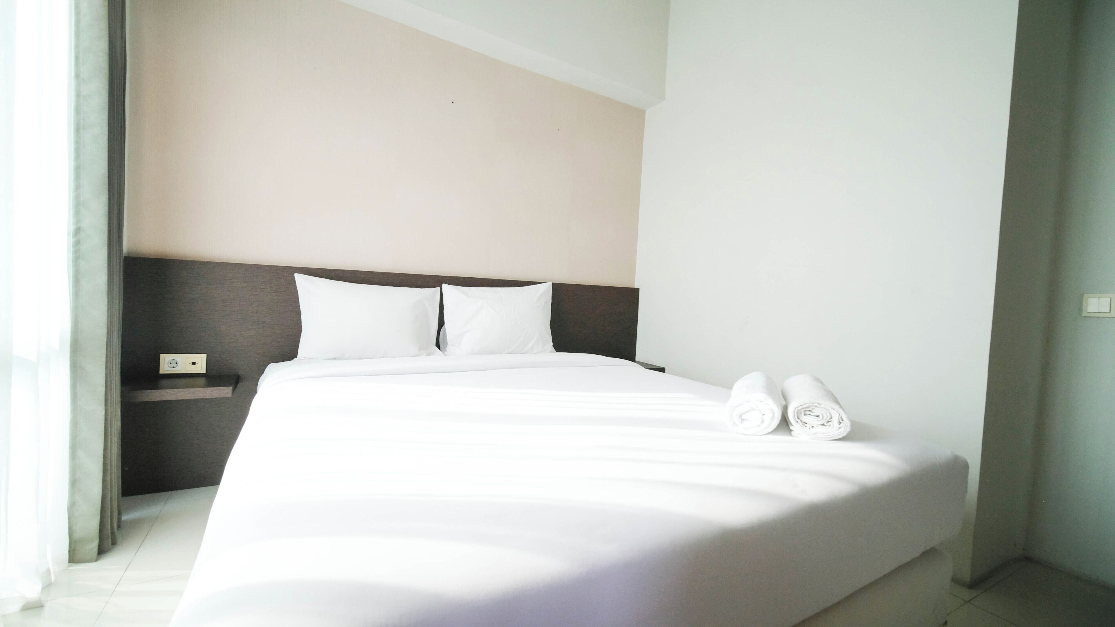 A neatly made bed with white linens and two rolled towels, a bedside table with a power outlet, and a plain wall with a light switch.