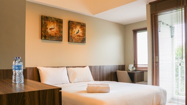 A hotel room with a large bed, two wall-mounted artworks, a wooden bedside table with a water bottle, and a window with blinds. - Simply Look Studio Room LRT City Jatibening Apartment (Bekasi)