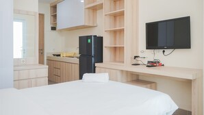 A modern hotel room with a bed, a flat-screen TV mounted on the wall, a built-in kitchen area with a refrigerator, and wooden shelving. - Cozy Stay And Good Deal Studio Apartment B Residence (Pagedangan)
