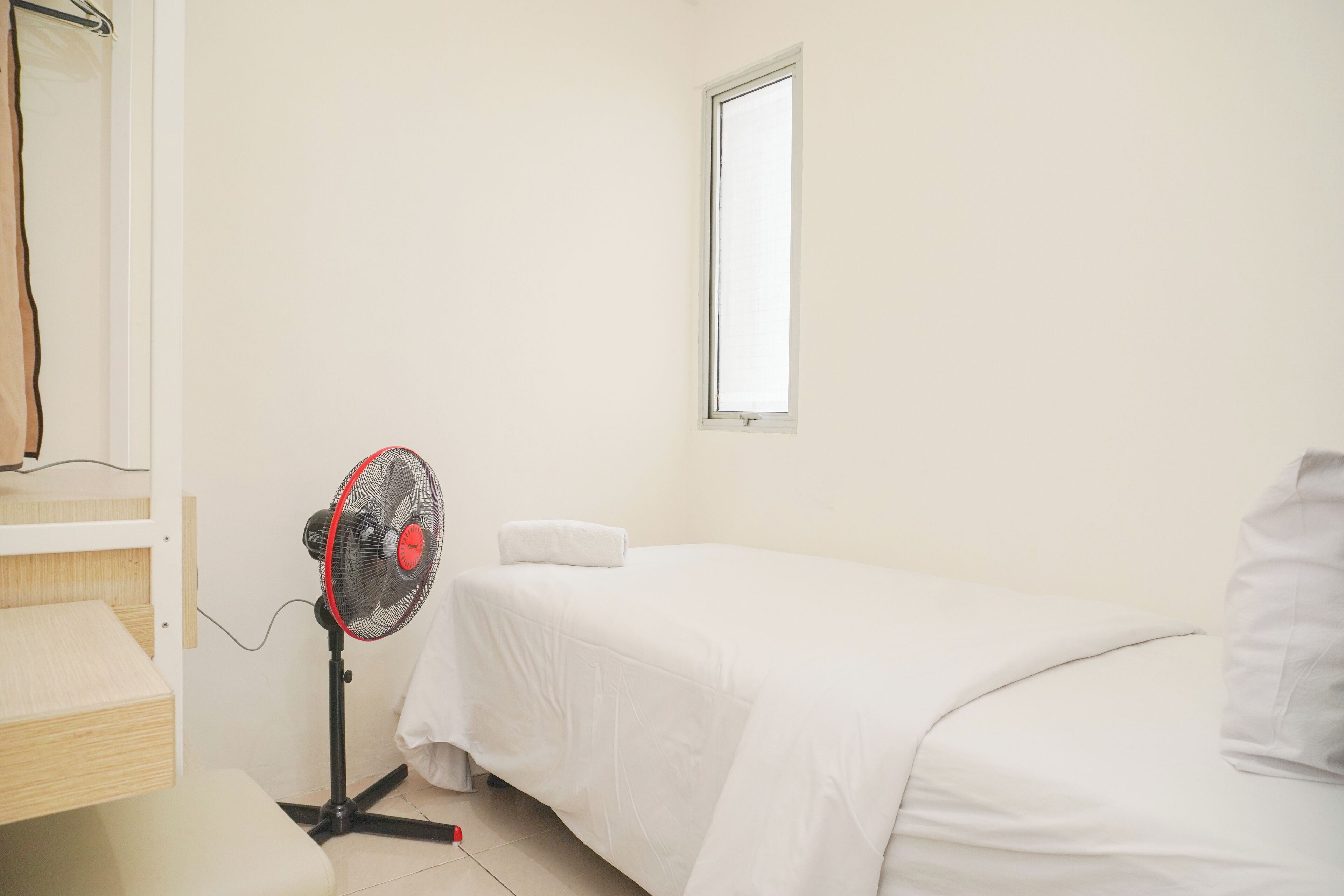 Ein kleines, sauberes Zimmer mit einem Einzelbett, einem Standventilator, einem Fenster und einem Holzregal.