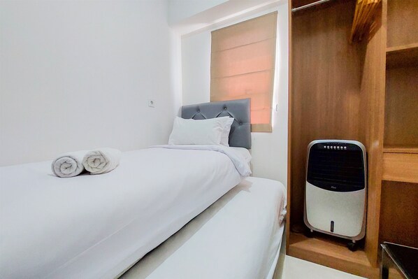 A neatly made bed with white linens and two rolled towels, a grey upholstered headboard, a window with blinds, and a portable heater.
