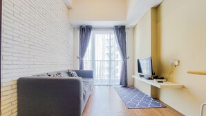 A modern living room with a grey sofa, a TV on a white shelf, and a view through a glass door. - Strategic And Restful 1Br At Casa De Parco Apartment (South Tangerang)