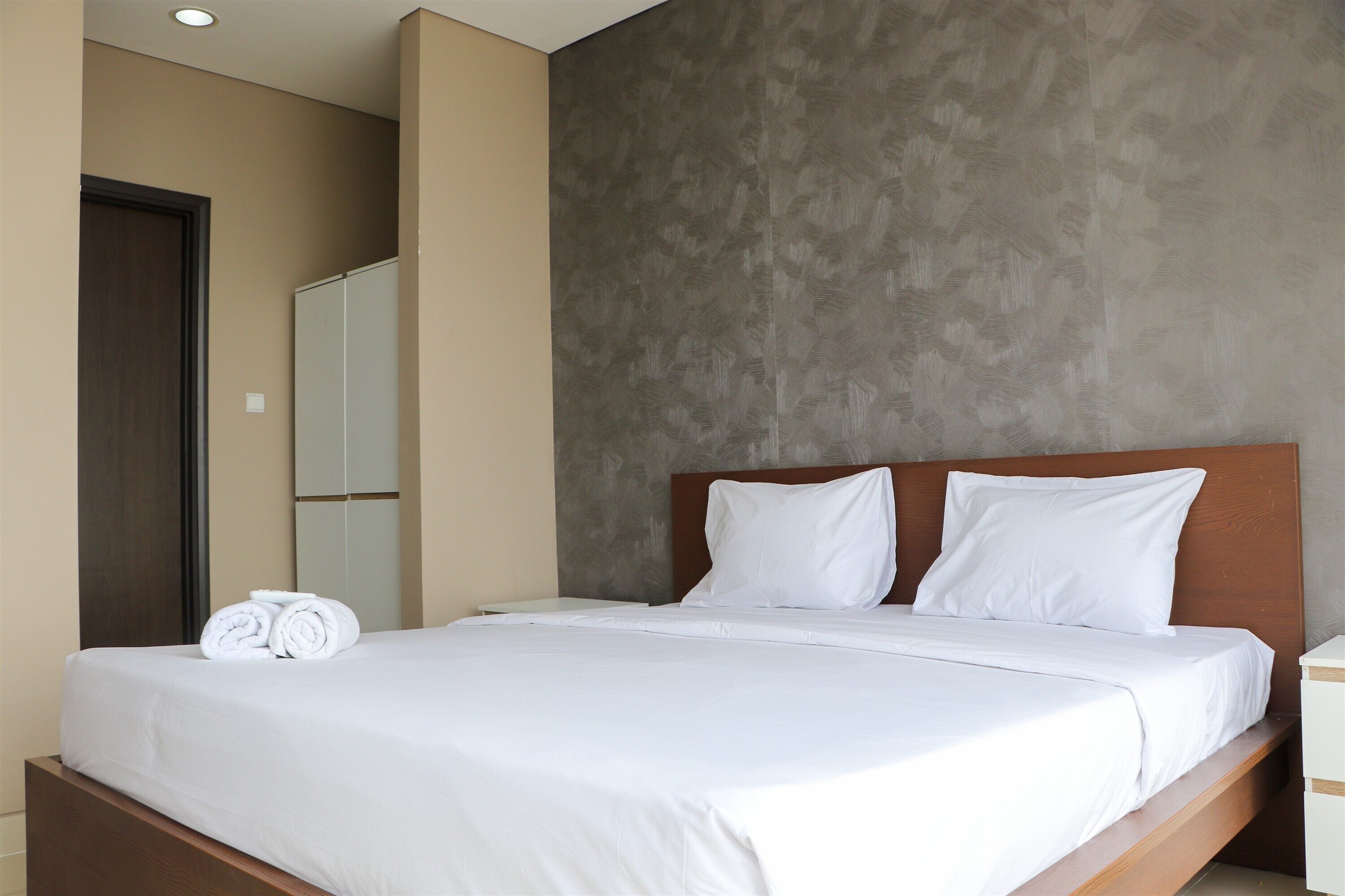 Un lit bien fait, avec du linge de lit blanc et des oreillers, une tête de lit en bois et un mur gris à la texture intéressante.