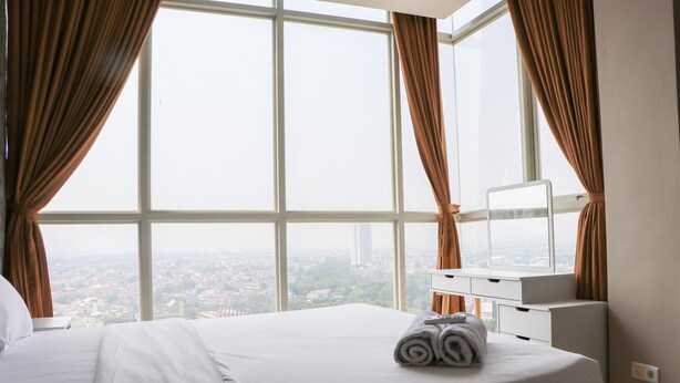 A hotel room with a large window, a bed with white linens, a bedside table with a lamp, and a view of a cityscape. - Spacious And Comfy 3Br Ciputra International Apartment (Jakarta)