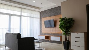 A modern living room with a grey sofa, a flat-screen TV mounted on a wooden panel, a potted plant, and a small table. - Spacious And Comfy 3Br Ciputra International Apartment (Jakarta)