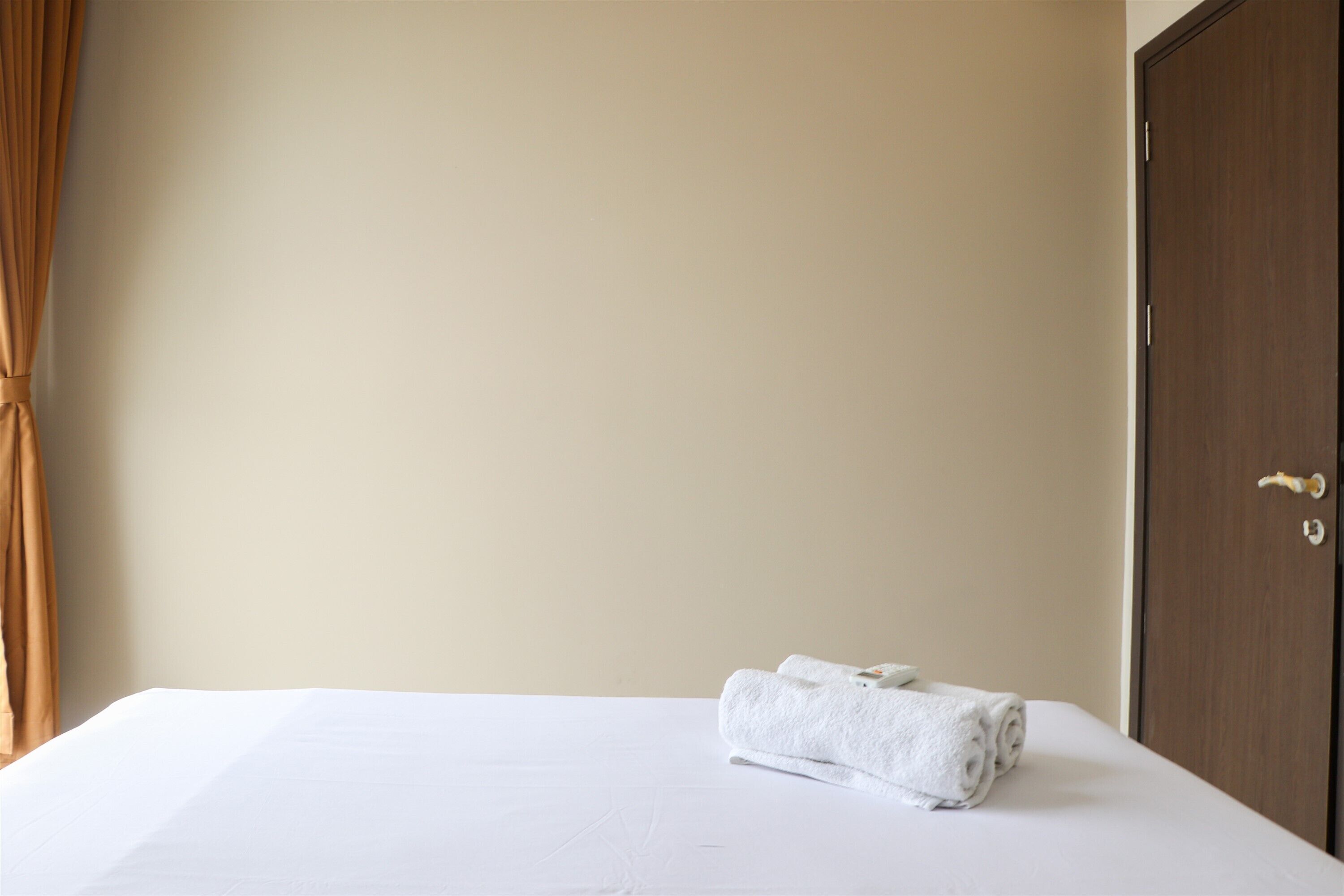 Una cama bien hecha con sábana blanca y una toalla blanca enrollada, una puerta marrón a la derecha y una pared color beige.