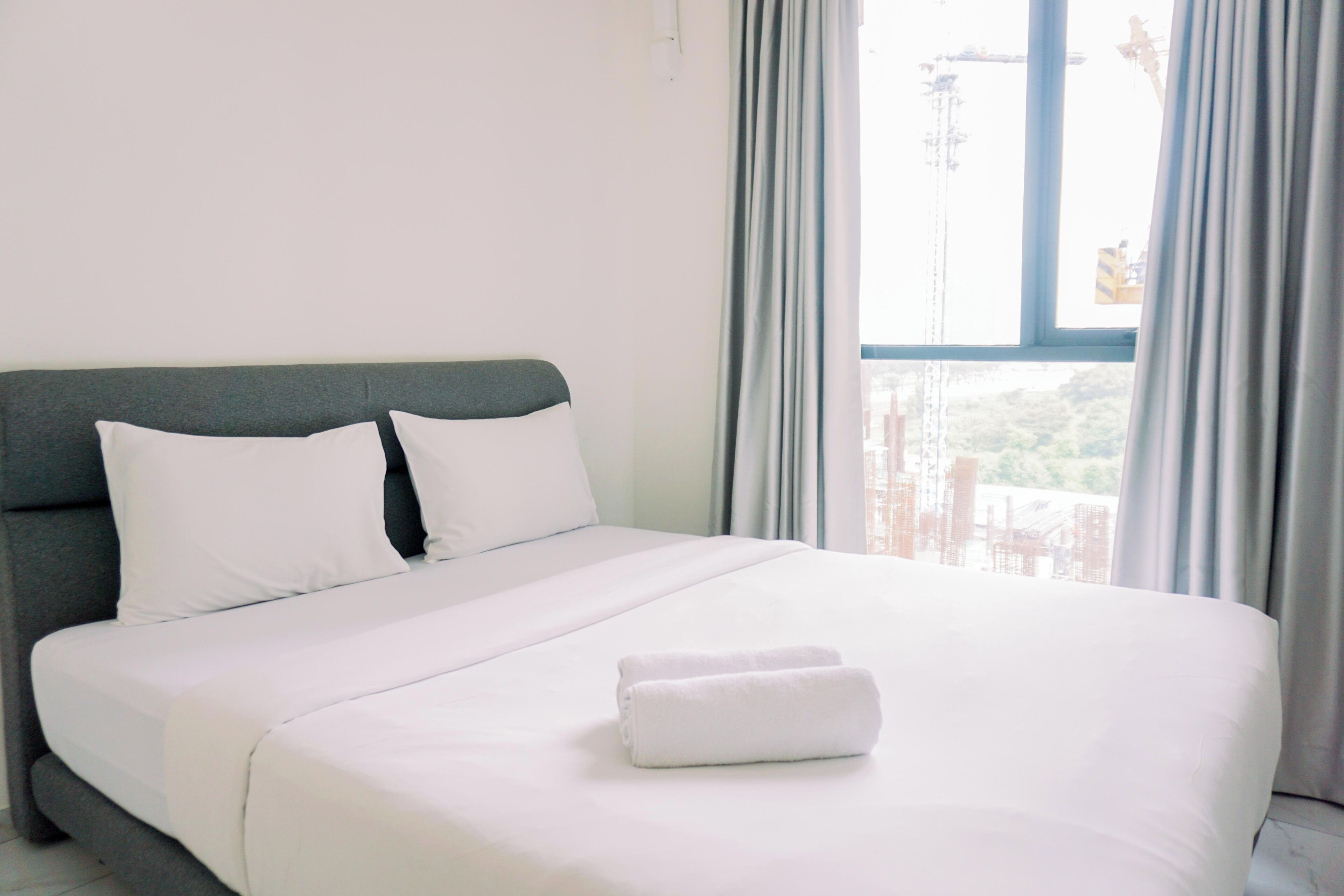 A neatly made bed with white linens and two rolled towels, a grey headboard, and a view of a construction site through a window with grey curtains.