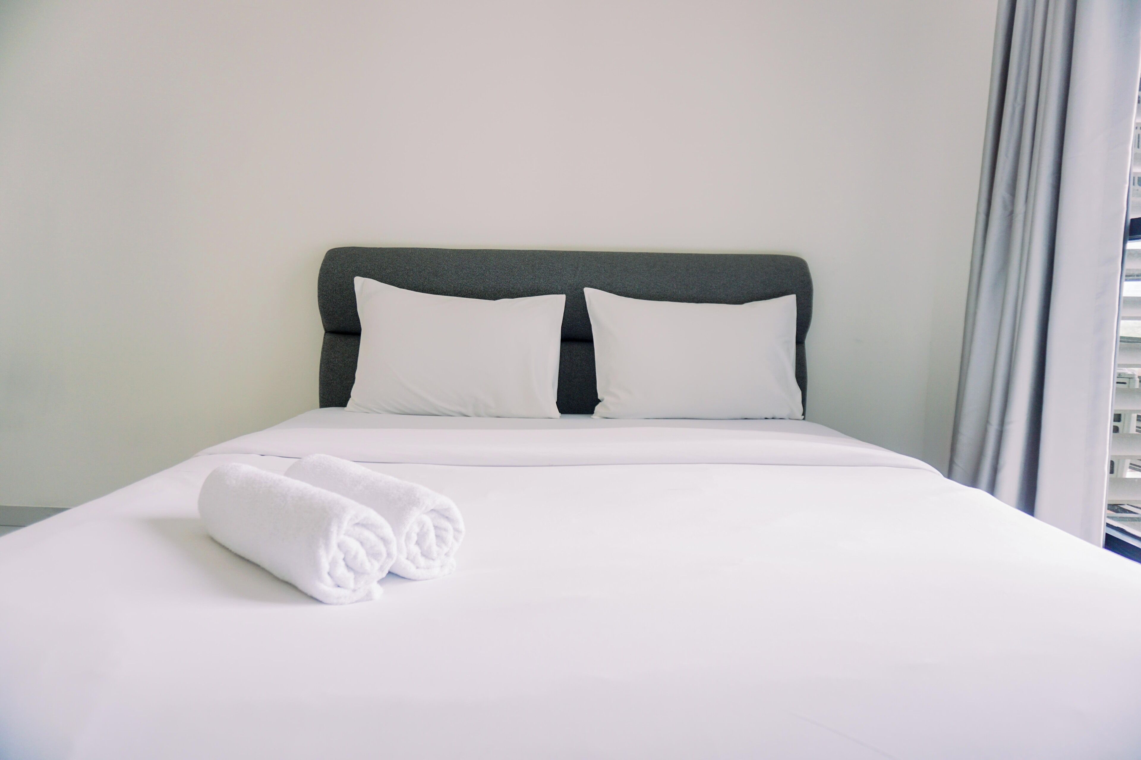 A neatly made bed with white linens and pillows, a rolled white towel, and a grey headboard.