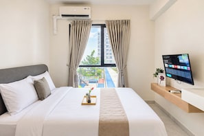 Ein Hotelzimmer mit Bett, Fernseher, Fenster mit Vorhängen und Blick auf ein Gebäude und Grünfläche.