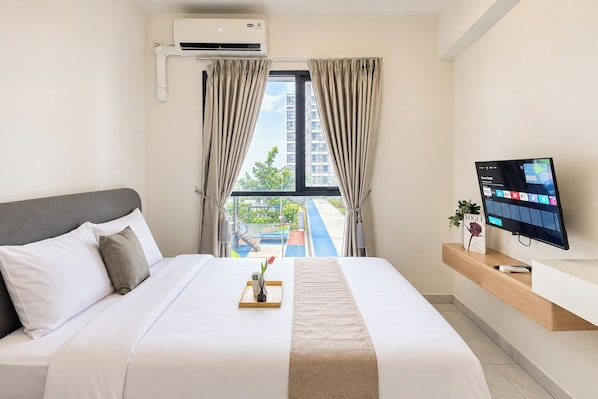 A hotel room with a bed, a TV, a window with curtains, and a view of a building and greenery outside.