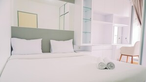 Una cama bien hecha con dos almohadas, un par de toallas enrolladas y una silla en una habitación luminosa con un ventanal grande y un espejo.