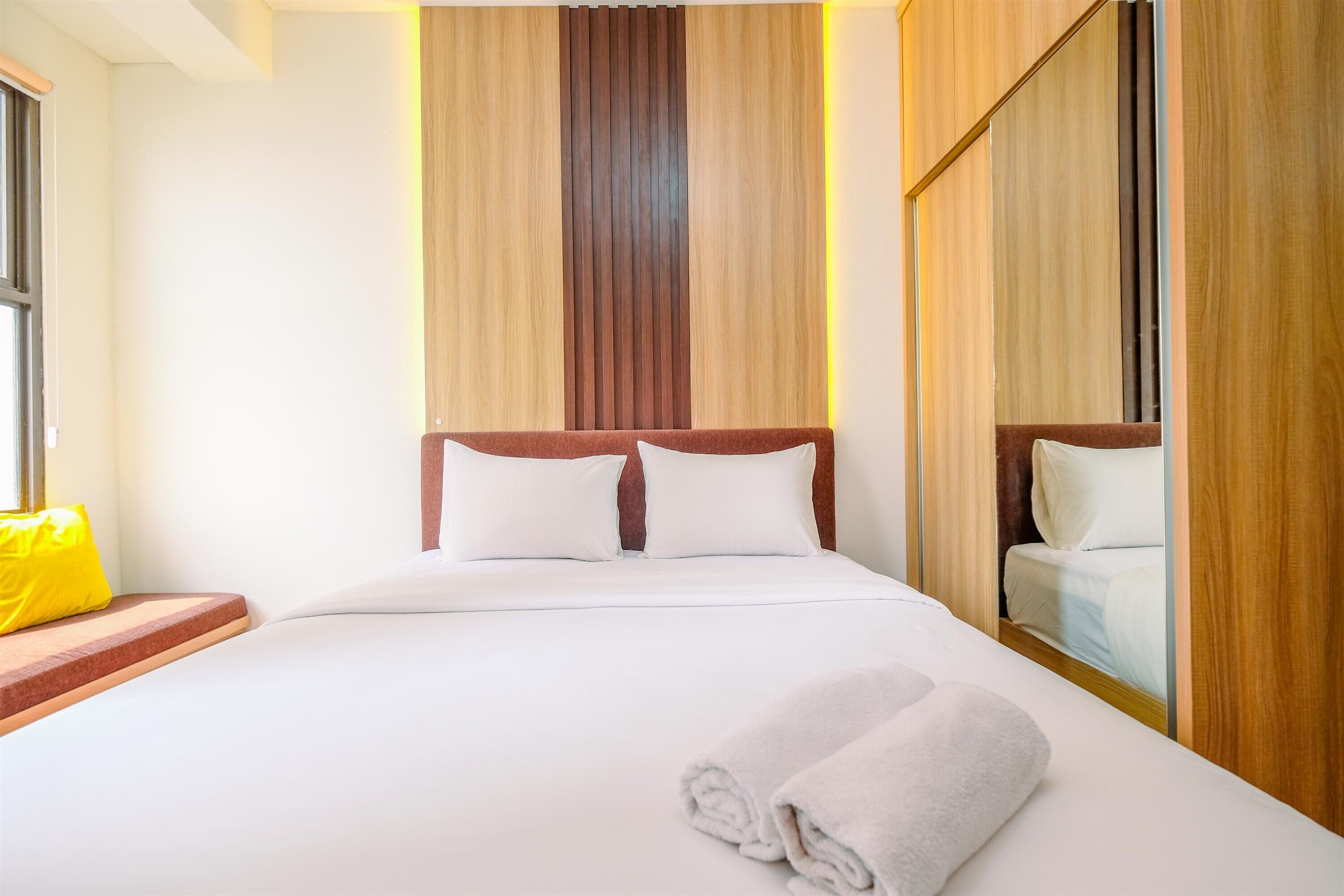 Una cama bien hecha con sábanas blancas y dos almohadas, un cabecero de madera y un cojín amarillo brillante sobre un banco de madera.