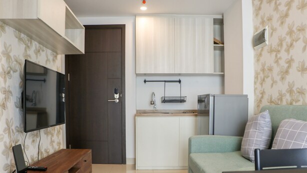 A compact living space with a kitchenette, a flat-screen TV mounted on the wall, a wooden side table, and a green sofa with patterned cushions.