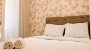 A neatly made bed with two pillows and rolled towels, a wooden headboard, and floral wallpaper.