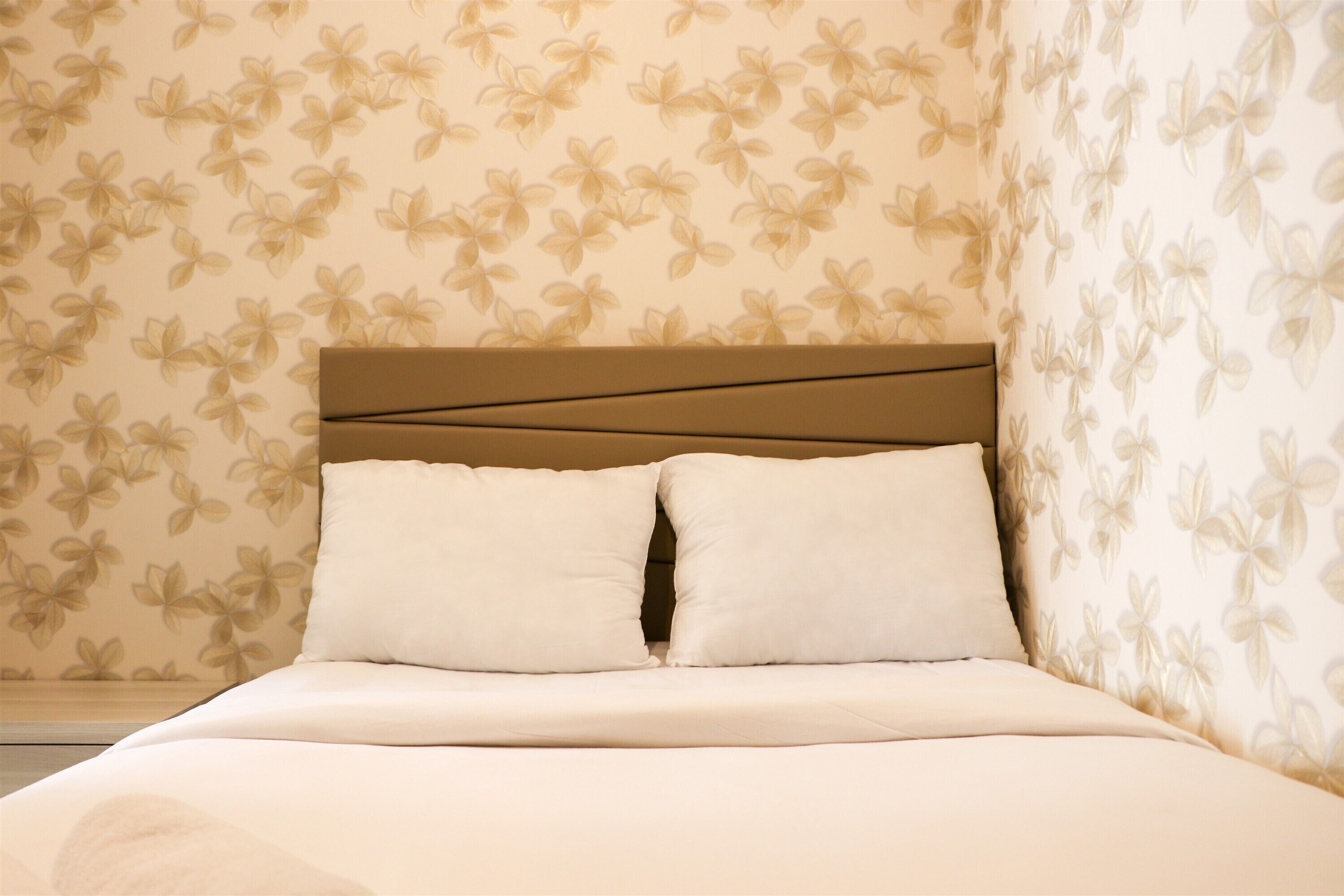 A neatly made bed with a headboard, a floral wallpaper pattern, and a nightstand to the left.