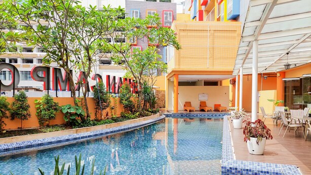 Una piscina con bordes de azulejos, rodeada de vegetación y un edificio con fachadas coloridas.