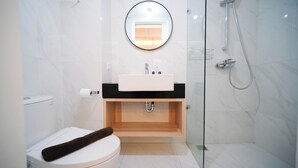 A modern bathroom with a white toilet, a wooden vanity, a round mirror, and a glass-enclosed shower area. - Cozy Studio Connected To Mall At The Vertu Apartment (Surabaya)