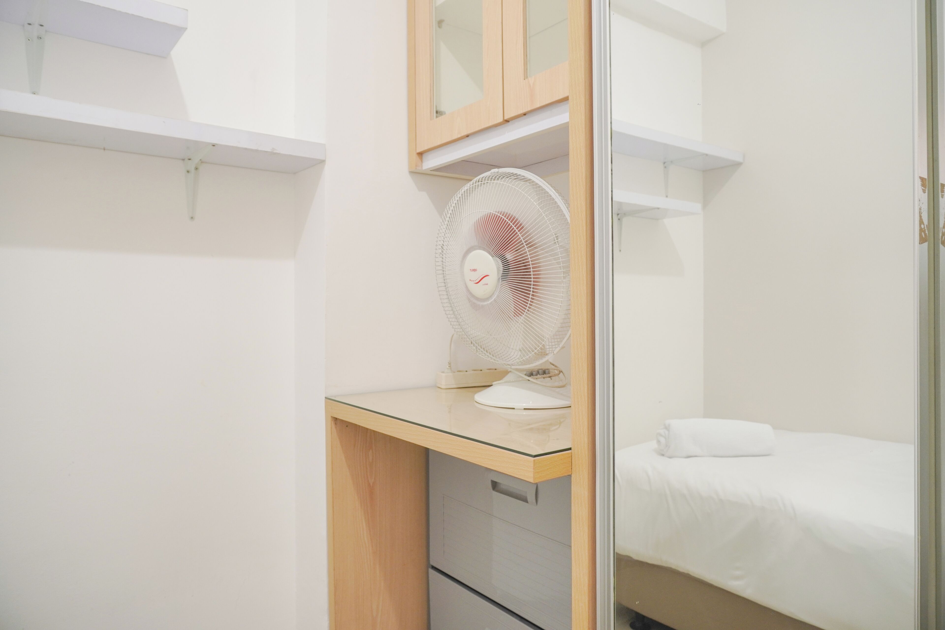 Un dormitorio pequeño y ordenado con un escritorio de madera, una cama blanca y un ventilador blanco.