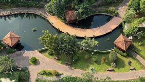 A park with a curved wooden bridge over a pond, surrounded by well-maintained greenery and two small pavilion structures.