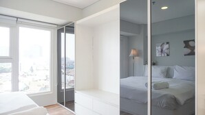 Un dormitorio moderno con un ventanal grande, una cama con ropa blanca, una mesita de noche y un espejo.