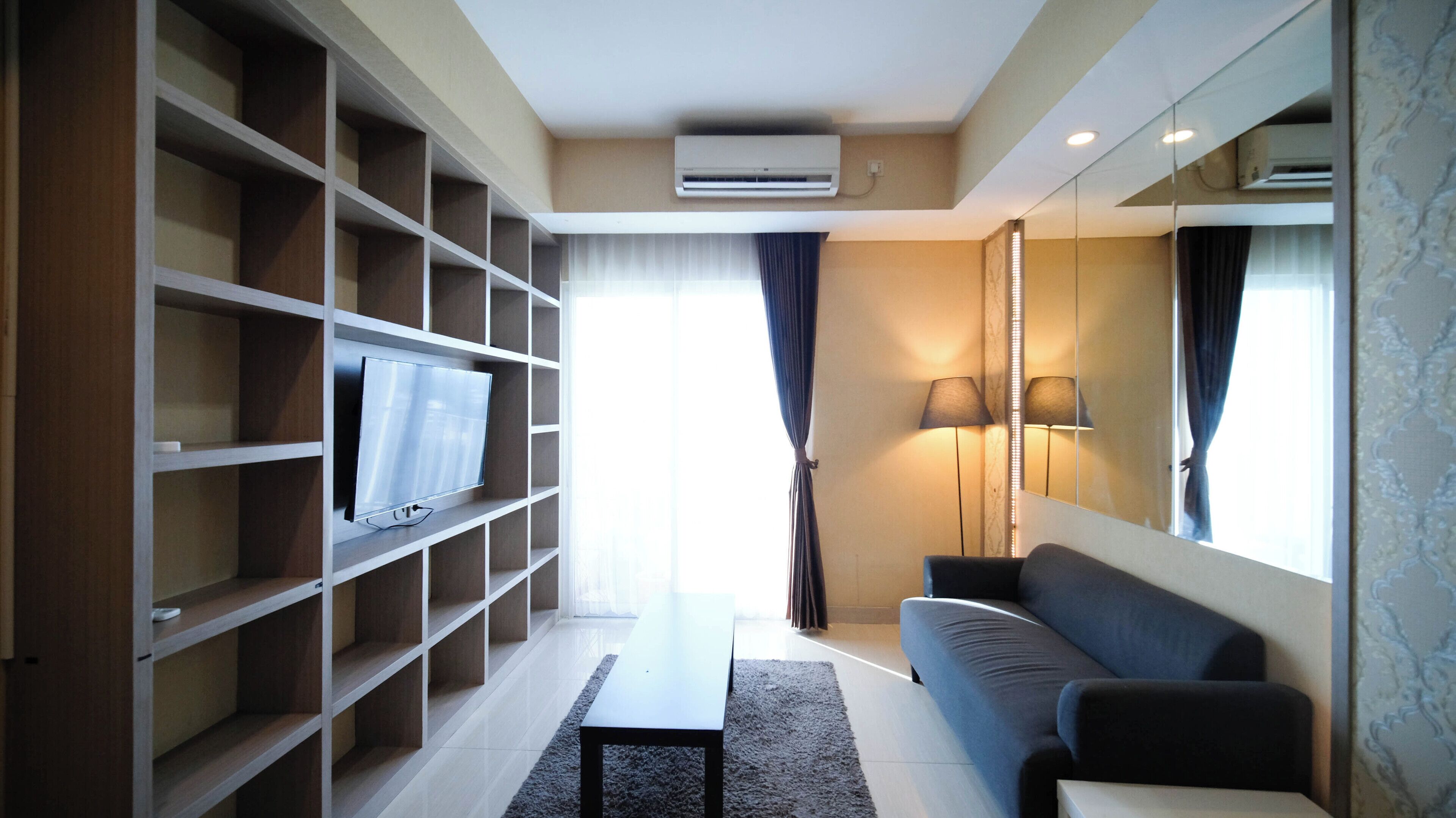 A modern living room with a flat-screen TV on a white shelf, a grey sofa, a small coffee table, and a large window with curtains.
