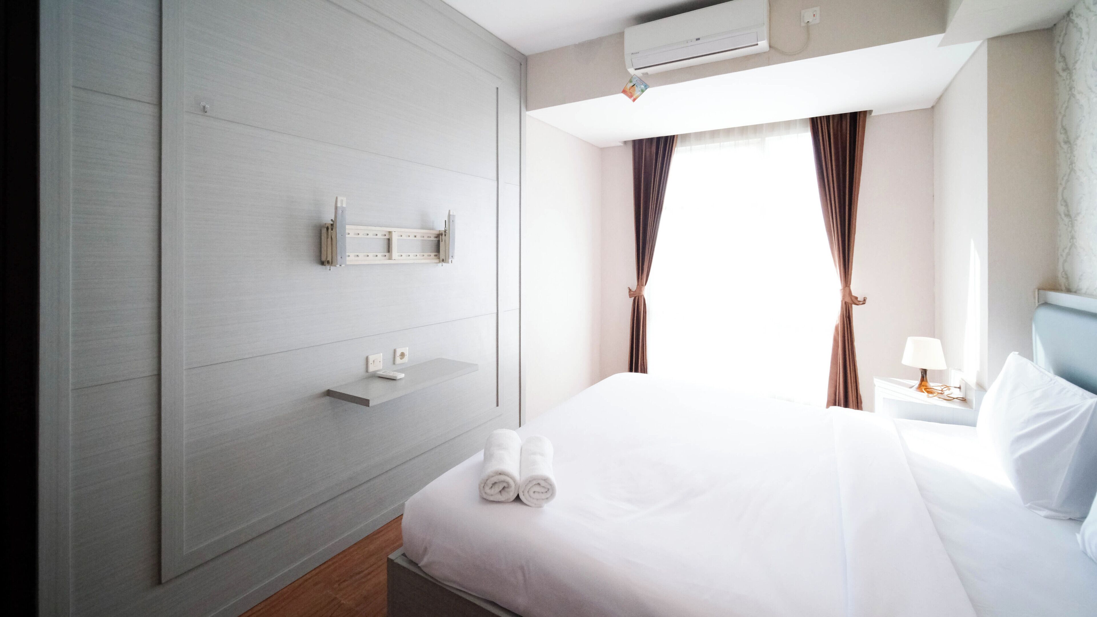 A hotel room with a bed, a nightstand, a wall-mounted shelf, and a window with curtains.